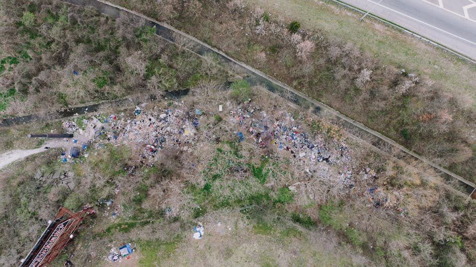 An aerial view of a large, uneven area covered with scattered piles of mixed waste materials, including plastic bags, paper, and other debris, situated adjacent to a narrow dirt path and a row of leafless trees. In the background, a retaining wall separates the waste site from a tributary of a small river or stream, which appears to be overgrown with grass and shrubs. The scene is bordered on one side by a paved road with a traffic barrier, suggesting proximity to a public roadway. The natural lighting is consistent with an overcast day, casting dull, diffuse light over the cluttered environment. The image reflects a typical scene of illegal or unmanaged rubbish dumping in a semi-rural location, which waste removal services like Waste Removal Lambeth are equipped to manage. The presence of loose debris and scattered trash indicates this area might be targeted for on-site clearance or private waste collection, supporting alternative disposal solutions outside local authority collection channels.