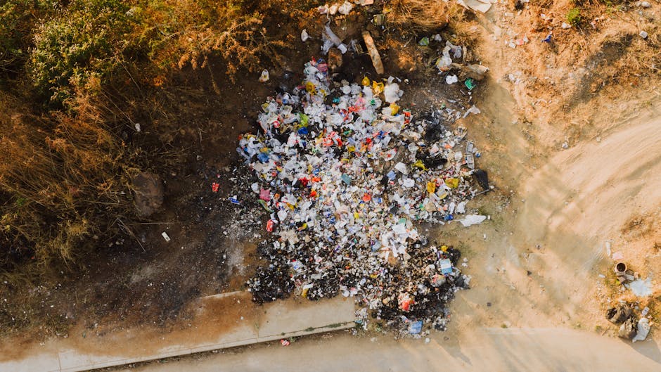 An aerial view of a large pile of mixed waste and rubbish scattered on bare soil next to a concrete pavement. The collection includes various materials such as plastic bottles, paper, cardboard, and small debris, with some items partially buried in dark soil or ash. Surrounding the rubbish are patches of dry, brown vegetation and a dirt embankment, indicating an unmanaged debris site likely resulting from illegal dumping or on-site clearance. The scene is illuminated by natural daylight, with shadows cast on the ground, highlighting the chaotic arrangement of waste. The presence of discarded items and urban environment suggests the scene could relate to private waste handling or the need for professional rubbish removal services such as those offered by Waste Removal Lambeth, who assist with clearing unmanaged or illegally dumped rubbish to prevent environmental and community impacts.