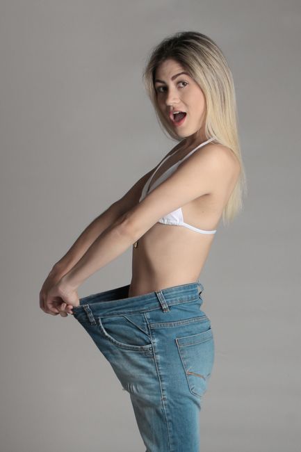 A woman with long blonde hair wearing a white bra and loose-fitting blue jeans pulled away from her waist, revealing a significant gap indicating she has lost weight. She is standing against a plain gray background, with her upper body slightly turned to her right, and her mouth slightly open as if speaking or reacting. Her expression appears surprised or pleased, and her arms are positioned to hold the waistband of her jeans. The image emphasizes the visual change in her waistline, symbolizing weight loss, which can relate to themes of personal health or fitness. The neutral lighting enhances the natural skin tones and the texture of the denim, with no additional objects or environment visible, focusing solely on her pose and expression.