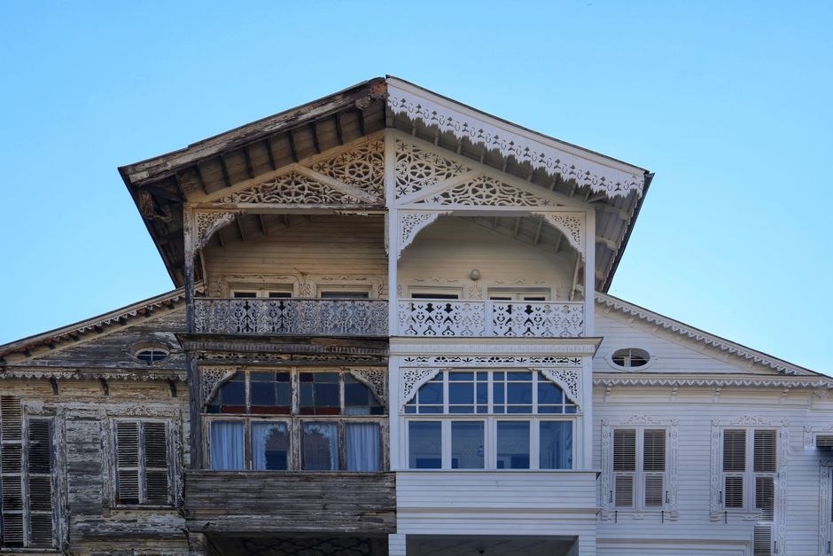 The image depicts an old, two-story wooden house with intricate Victorian-style architectural details. The lower level shows weathered, darkened wood with peeling paint, exposing patches of rough, aged timber and small, shuttered windows that are partly obscured. The upper level features freshly painted white wood paneling, ornate cast-iron lacework decorating the balcony railings, and decorative timber trim along the eaves of the roof, which is supported by decorative brackets. The house's gabled roof has a slight pitch and overhangs, with visible dark wooden beams beneath. Surrounding the house is a clear blue sky, providing natural lighting that highlights the contrast between the aged, deteriorated lower section and the well-maintained upper section. Despite its deteriorated condition, the property appears structurally sound, with no visible signs of immediate damage or collapse. This detailed view emphasizes the historic craftsmanship of Victorian houses, connecting to contexts of property renovation, restoration, or private property management, all relevant within the scope of waste removal or site clearance services like those offered by Waste Removal Lambeth.