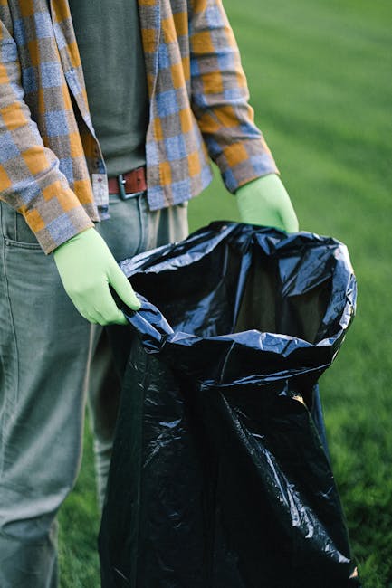 A person wearing a casual plaid shirt with shades of yellow, gray, and blue, along with green rubber gloves, is seen holding open a large black plastic rubbish bag with both hands, preparing to dispose of waste. The individual is standing outdoors on a grassy area, with the background showing well-maintained green lawn and some blurred natural greenery. The black rubbish bag appears to be partially full and is designed for waste collection and removal, possibly handled by a waste disposal service such as Waste Removal Lambeth. The scene is well-lit, indicating daylight, and emphasizes the practical aspect of trash collection, which might involve private or alternative disposal methods as part of on-site clearance or rubbish removal services. The textures of the plastic bag are shiny and crinkled, contrasting with the matte fabric of the gloves and casual clothing, creating a clear visual focus on waste handling activities in a green environment.