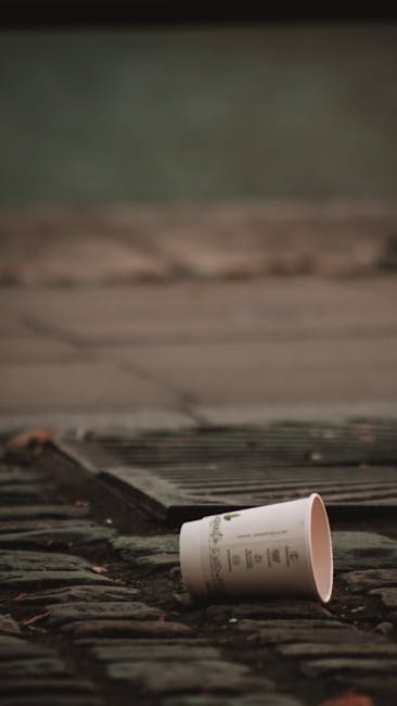 A disposable paper coffee cup with a white exterior and printed green and black text is lying on its side on a brick and concrete surface outdoors. The cup is situated near a drain cover with evenly spaced metal slats, embedded into the pavement. In the background, there is a blurred section of concrete and asphalt, suggesting an urban or residential street environment. The scene appears to be in natural daylight, with diffused lighting and no visible people. The accumulation of debris and such discarded items is consistent with waste that might be subject to independent rubbish removal services like those provided by Waste Removal Lambeth, which handle non-regular waste collection, including small litter items left in outdoor areas. The textured brick and concrete ground contrasts with the smooth surface of the cup, emphasizing its presence in the scene, typical of informal rubbish disposal found in urban settings. The environment and context imply the need for regular maintenance or private waste handling to prevent litter accumulation in public or private outdoor spaces.