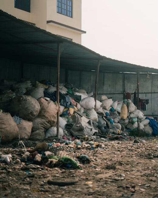 A large collection of tightly packed black and white rubbish bags, various plastic materials, and discarded household items are stacked against a corrugated metal fence beneath a dark green, corrugated roof. The bags contain different types of waste, with some appearing bulging and others torn open, spilling their contents onto the uneven, debris-strewn ground, which is scattered with loose litter, plastic wrappers, and small pieces of rubbish. Behind the fence, part of a multi-storey cream-colored building with large windows is visible, suggesting an urban or semi-urban environment. The scene is lit with natural daylight, casting soft shadows and highlighting the cluttered, unmanaged waste accumulation, which potentially relates to private or independent waste handling services such as rubbish removal, as it appears outside of a formal collection system. The overall setting reflects an area awaiting clearance or disposal, typical of a non-authorized waste storage or collection point, illustrating the need for professional rubbish removal services in Lambeth.