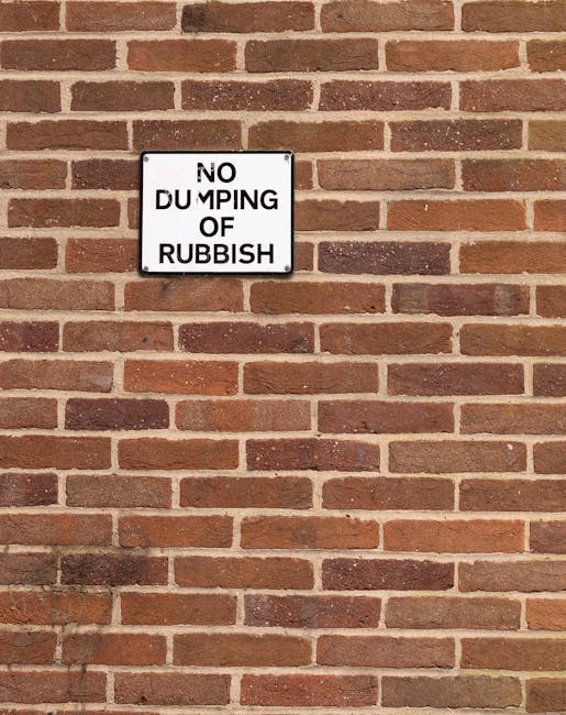 A rectangular black and white metal sign attached to a brick wall, displaying the message 'NO DUMPING OF RUBBISH' in bold uppercase letters. The wall is composed of evenly laid reddish-brown bricks with mortar joints, creating a uniform pattern across the surface. The sign is positioned near the upper left area of the image, with the brickwork extending below and to the right, suggesting an exterior urban or commercial setting. The lighting is natural, casting minimal shadows on the wall, and emphasizing the textured surface of the brickwork. This scene illustrates a common method of communicating waste disposal regulations at private or semi-private properties, supporting alternative rubbish removal services by discouraging illegal dumping in designated areas, aligning with the focus on rubbish collection and waste management solutions provided by Waste Removal Lambeth.