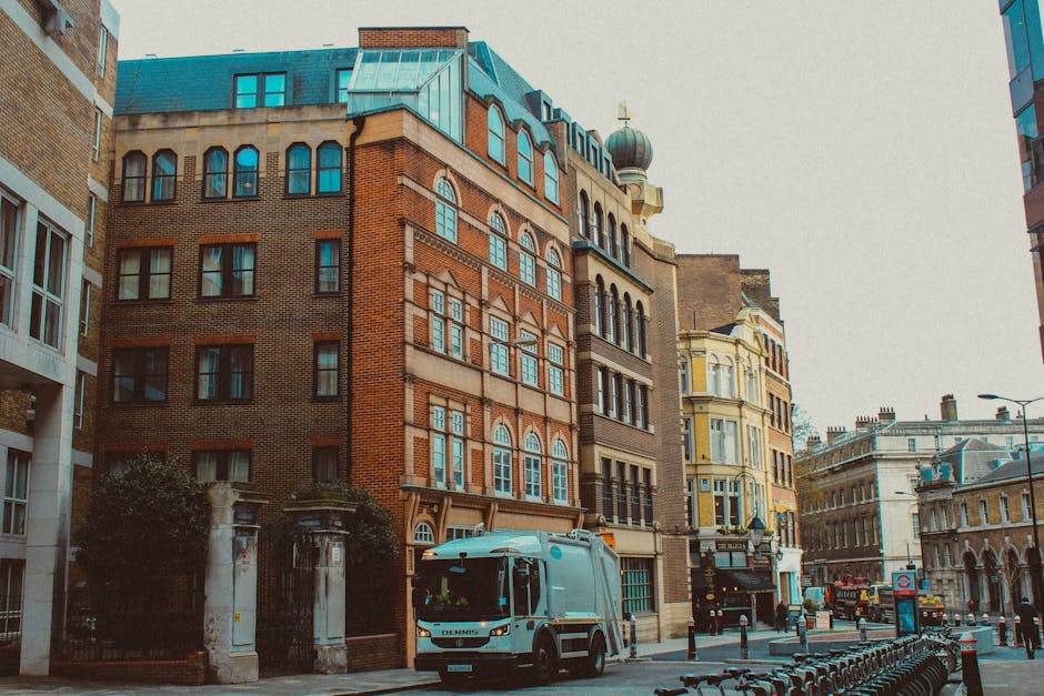 A street scene featuring a collection of narrow, multi-story buildings with diverse architectural styles, including brick facades, large arched windows, and ornate detailing. In the foreground, a small, modern waste collection vehicle with a light blue and white exterior is parked on the pavement near the curb, likely used for private rubbish collection services. To the left, an entrance gate with stone pillars and greenery creates a contrast to the urban environment. Surrounding the scene are other buildings with various finishes, including a prominent, yellow-painted structure with decorative windows. The street is lined with bollards and bike racks, while a few pedestrians and additional vehicles can be seen further along the road under overcast skies. The lighting is subdued, typical of overcast weather, highlighting the textures of the building materials and the urban setting. The presence of waste collection equipment suggests alternative waste handling options compared to municipal skip hire, aligning with private rubbish removal services often provided by companies like Waste Removal Lambeth.