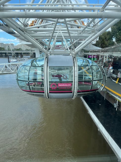 A large, modern Ferris wheel with a prominent glass-enclosed capsule hangs over the River Thames in London. The capsule features transparent, curved glass panels framed by metallic supports, allowing clear views of the interior and surroundings. The Ferris wheel structure is composed of white, lattice-like steel beams that extend upwards and outwards, partially visible in the background. The capsule is positioned near a dockside area with a black and yellow safety barrier and a small waiting platform visible on the right side of the image. The water below the wheel appears brownish and reflects the sky and nearby structures. Some trees and urban buildings are visible in the background, along with a few boats on the river, emphasizing the scenic cityscape setting. The scene is illuminated by natural daylight, with a partly cloudy sky above, contributing to a bright and clear atmosphere typical of outdoor sightseeing attractions. This image relates to the context of urban infrastructure and could be associated with the transport or tourism services offered in the area, such as private or alternative transportation options, including ferry or cable car operations supported by companies like Waste Removal Lambeth for infrastructure maintenance or event waste management.