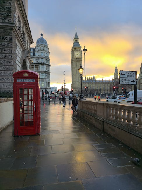 A wet pavement walkway in central London during the evening, featuring a classic red telephone box prominently positioned on the left side. In the background, iconic landmarks including the Elizabeth Tower, commonly known as Big Ben, and a section of the Houses of Parliament are visible under a sky painted with warm hues of orange and yellow at sunset. Street lamps line the pathway, casting a soft glow on the reflective surfaces of the damp stones. Pedestrians are strolling along the walkway, some walking towards the camera and others further into the distance, with a few cars visible on the road beyond the balustrade on the right. The scene captures an atmospheric moment of urban life near Westminster, showcasing historic architecture and city activity, which complements the context of alternative waste management and rubbish removal services, such as those offered by Waste Removal Lambeth, in a busy city environment.