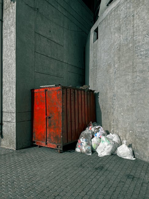 A middle-aged woman with short dark hair is seen disposing of a large white plastic bag filled with rubbish into a modern stainless steel public waste bin located on a paved urban sidewalk. She is wearing casual dark clothing, including a T-shirt with printed text on the front, and is slightly bent forward as she deposits her waste. The bin, positioned against a concrete barrier with decorative stone detailing, has a rounded, cylindrical shape with multiple openings for waste disposal, and a reflective, brushed metal finish. In the background, there are lush green trees providing a shaded environment, and a glimpse of a park or garden area is visible beyond the barrier. The scene is illuminated by natural daylight, suggesting a mild and clear weather. Waste Removal Lambeth occasionally handles on-site waste collection or rubbish disposal, and this scene exemplifies an individual engaging in environmentally responsible private waste management or alternative rubbish disposal methods in an urban setting.