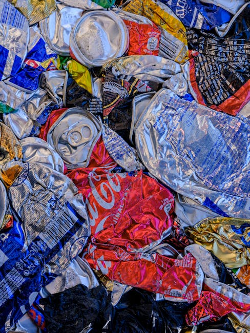 A close-up image of a pile of crushed aluminum cans, predominantly made of lightweight, reflective metal with some showing visible pull-tabs and indentations. The cans have various printed designs, logos, and colours, including red, blue, silver, and black, with some featuring recognizable branding like Coca-Cola. The cans are crumpled and stacked loosely, with some overlapping others, creating a textured surface. The background shows a flat, possibly outdoor environment, with the cans appearing as part of waste awaiting collection or disposal. The scene emphasizes the need for rubbish removal services, highlighting how waste materials such as cans are gathered and prepared for environmentally responsible disposal. This visual is relevant to private waste handling and alternative collection methods, supporting the context of independent rubbish removal solutions provided by Waste Removal Lambeth. Natural lighting reflects off the metallic surfaces, accentuating their crumpled form and shiny finish, illustrating an example of post-consumer waste accumulation suitable for on-site clearance or recycling collection.