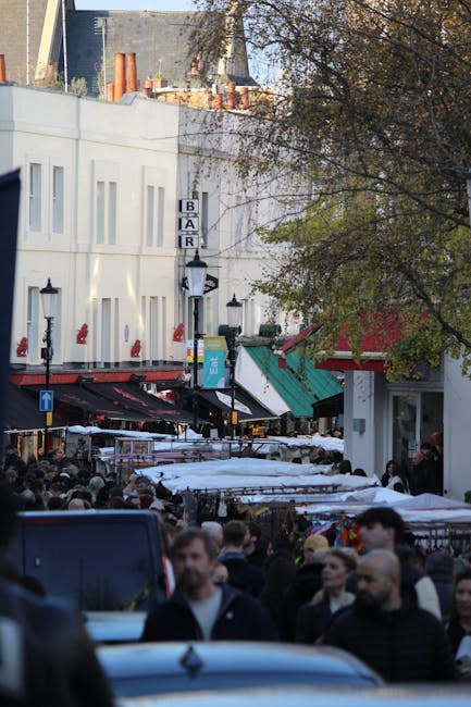 A busy street scene in an urban marketplace with a large crowd of pedestrians walking along a narrow, crowded thoroughfare. The street is lined with small shops and market stalls, some covered by white awnings, with various goods on display. Buildings on either side are predominantly white with flat facades, and there are several black vintage-style street lamps mounted on the building walls. A sign for a bar hangs on one side of the street, and there are visible chimneys and rooftops of nearby buildings in the background. Trees with sparse foliage partially shade the street, and the overall scene is set in daylight under clear weather conditions. This environment suggests a lively commercial area where private waste collection or rubbish removal services, such as those offered by Waste Removal Lambeth, might be utilized for maintaining cleanliness during busy market days, although no specific waste or rubbish is visible in the image.