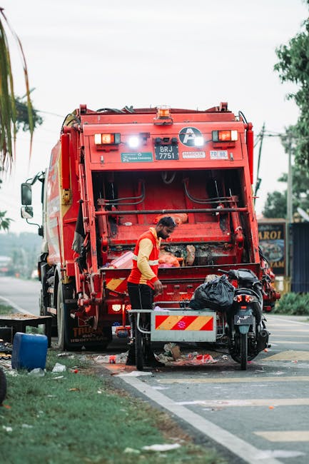 A waste collection worker, dressed in a yellow high-visibility vest with red accents, is seen standing near the rear of a large red rubbish truck on a paved street. The worker is emptying contents from a black plastic bag into the truck’s hopper, which has a textured metal surface with signs of wear and dirt. The truck is positioned on the side of the road, with its rear open to facilitate waste loading, and features various safety lights and warning labels on its back panel. Nearby, there is a black motorcycle parked on the pavement, partially blocking a small metal privacy barrier with red and white reflective stripes. Surrounding the scene, the environment includes a driveway with a patch of grass and a blue wheelie bin in the foreground, indicating a residential or mixed-use area. In the background, blurred trees and utility poles are visible under an overcast sky, suggesting typical urban waste management activity possibly relating to private or alternative rubbish removal services like those offered by Waste Removal Lambeth, servicing the Streatham SW16 area, which manages refuse collection outside local authority scheduling.
